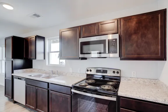 A Cypress Point kitchen with dark cabinets, a refrigerator, an electric range with a microwave above it, a dishwasher, a double sink and light counters.