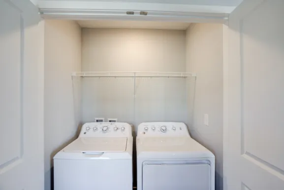 The laundry space inside a Cypress Point apartment, with a top-loading washing machine and a dryer standing side by side beneath a wire shelf.